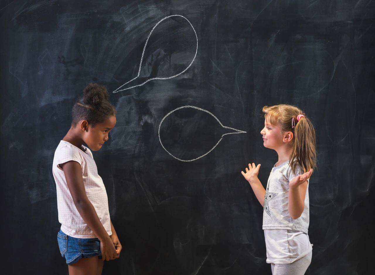 Two elementary school students speak and gesture to each other in a contentious conversation in front of a chalkboard with chalk speech bubbles drawn over each.