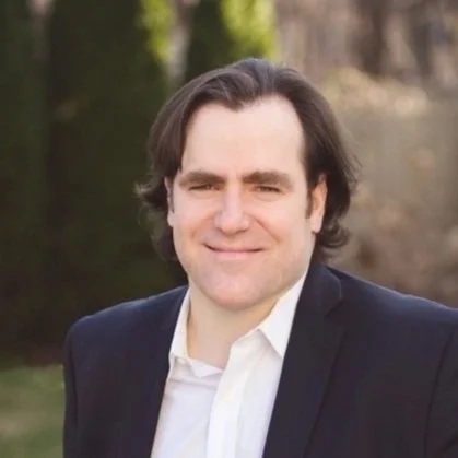 A person wearing a dark blazer and a white collared shirt, standing outdoors with greenery and soft natural light in the background.