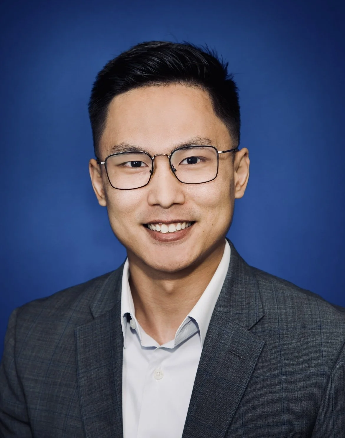 A person dressed in a gray plaid suit jacket and a light collared shirt, posed against a bright blue background.
