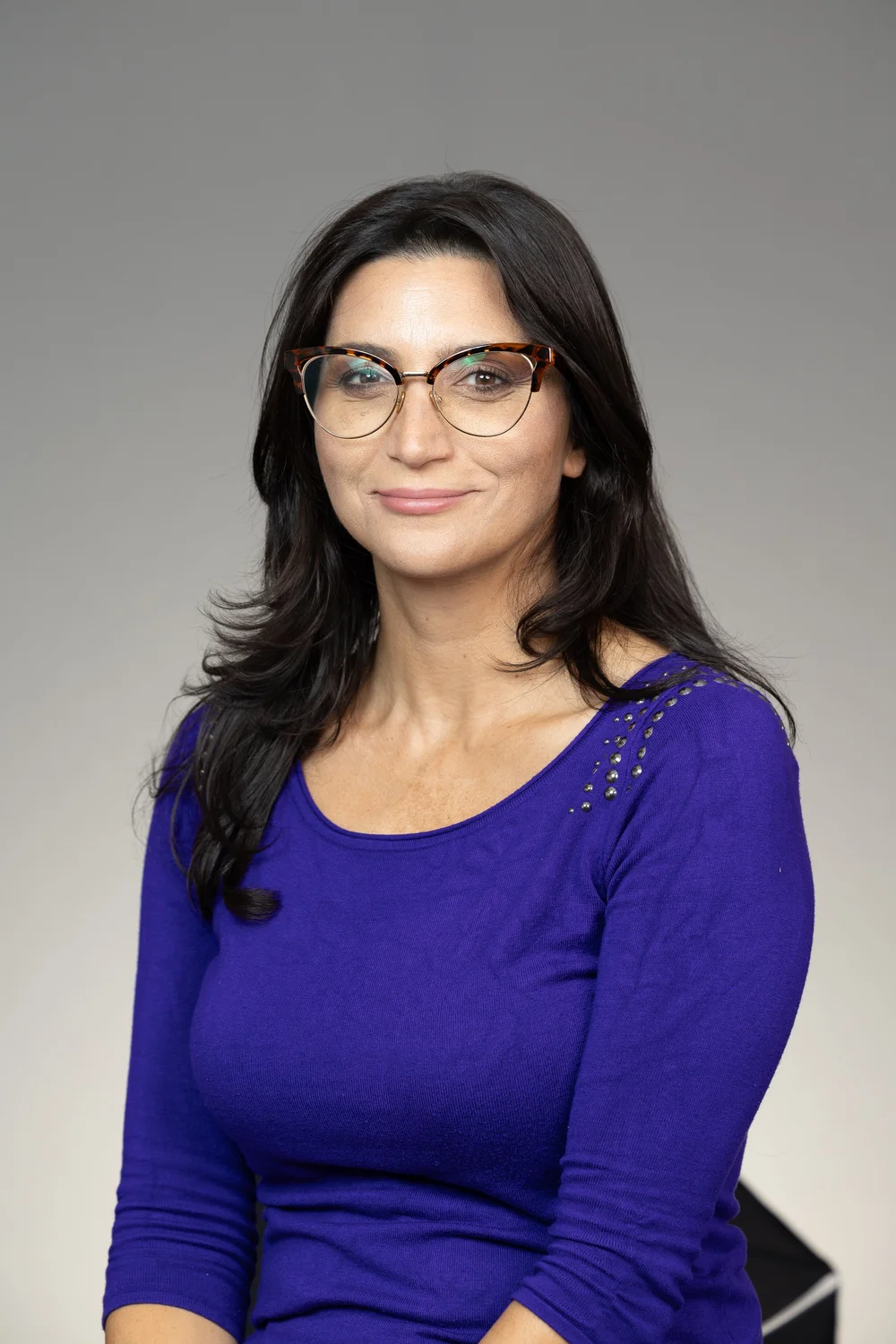 A person wearing a royal blue long‑sleeve top with decorative studs on the shoulder, seated against a light gray background.