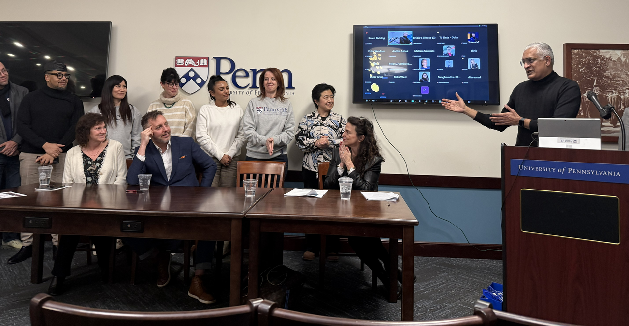 Raghu stands at a podium with a zoom screen of faces behind him and a collection of eight CLO students seated and standing at a table next to him in front of a Penn logo