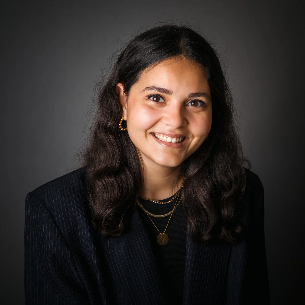 A person wearing layered gold necklaces, gold hoop earrings, and a dark pinstripe jacket, posed against a dark studio backdrop.