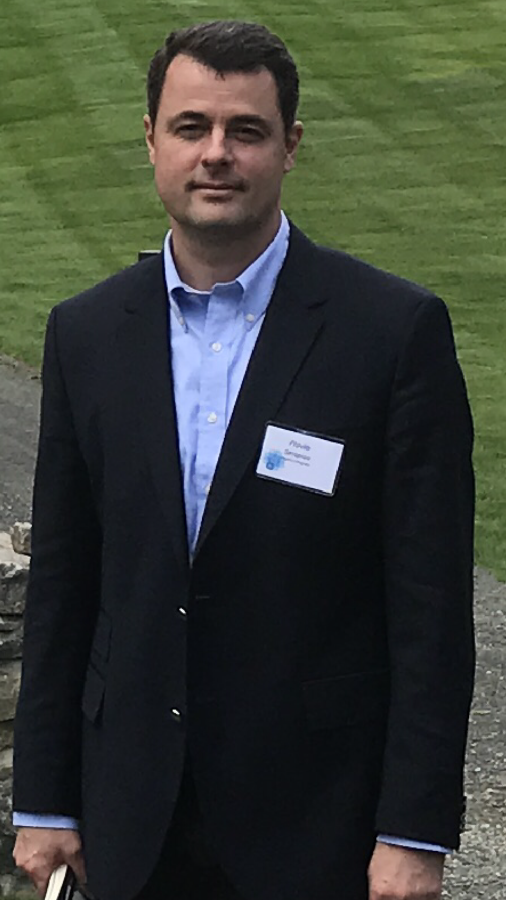 A person wearing a dark jacket and light blue collared shirt, standing outdoors near a grassy area and stone walkway. A name badge is clipped to the jacket.