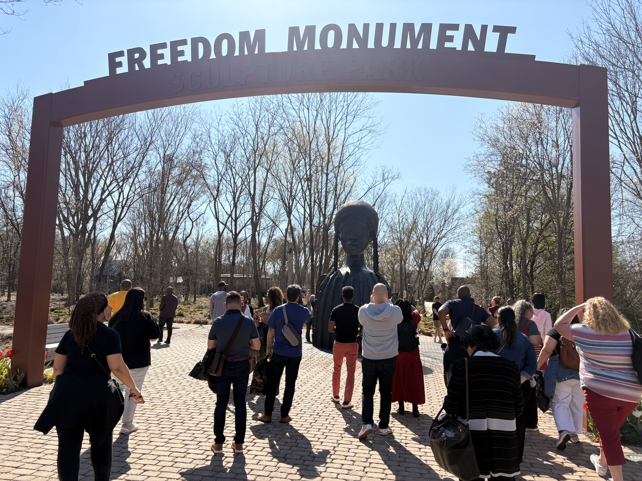A group of attendees walk under a sign for the Freedom Monument Sculpture Garden on a sunny day towards a large bronze sculpture of a Black woman with braids