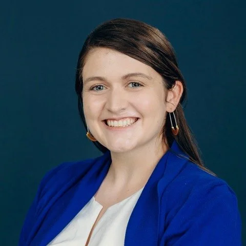 A person wearing a bright blue blazer over a white top, shown from the torso up against a dark teal background.