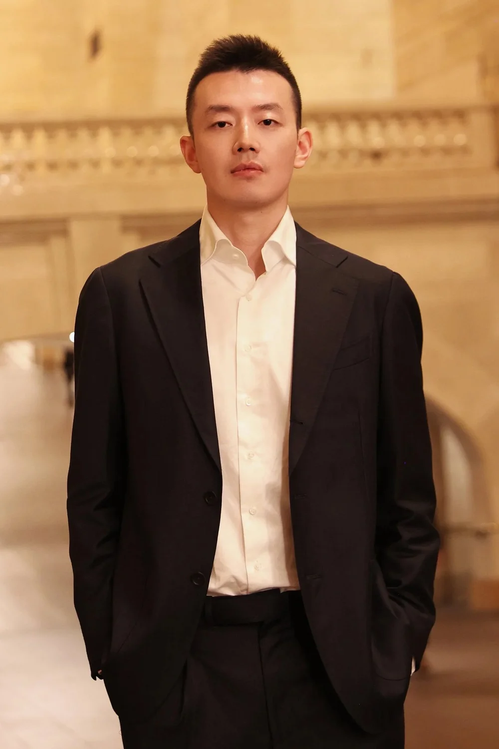 A person dressed in a dark suit with a white collared shirt, standing indoors in a large, warmly lit architectural space.