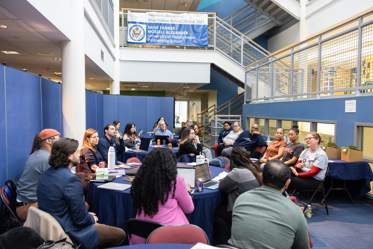 A group of SLP students gather around tables talking to Penn Alexander staff under a sign that reads "Sadie Tanner Mossell Alexander: University of Pennsylvania Partnership School"