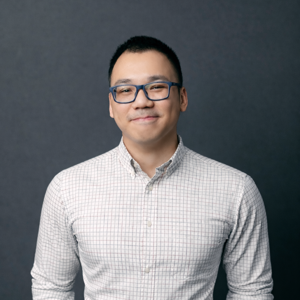 Person wearing a light-colored button‑down shirt with sleeves rolled up, standing against a dark gray studio background