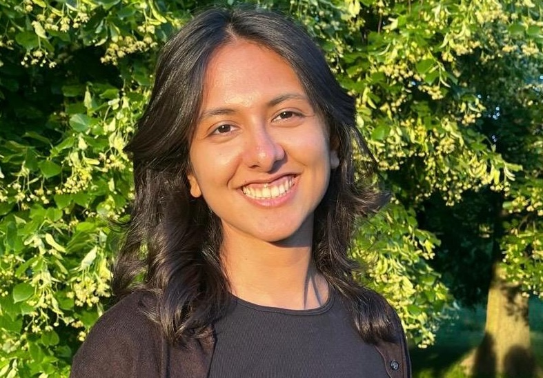 Person with shoulder‑length dark hair wearing a black top, standing outdoors in front of leafy green trees in natural sunlight