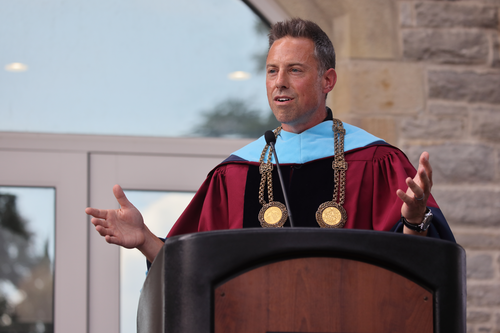 President vandenBerg wears graduation regalia and speaks at a podium outdoors. 
