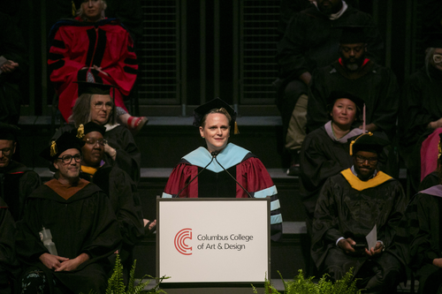 President Corn wears academic regalia and speaks in front of a crowd in academic regalia at a podium with the name and logo of Columbus College of Art and Design on it. 