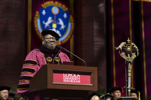 Dr. Cole wears graduation regalia and speaks at a podium that features a sign saying Loyola University New Orleans
