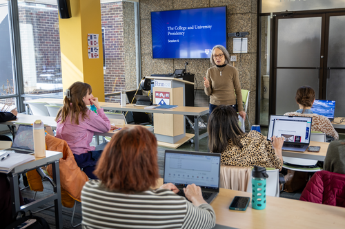 Julie Wollman stands in front of a class with a screen behind her with text on it that reads "The College and University Presidency"