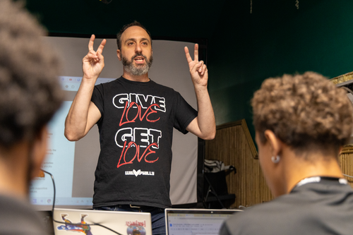 Cory makes air quotes with his fingers, photographed from behind a row of students in his class, wearing a We Love Philly t-shirt. 