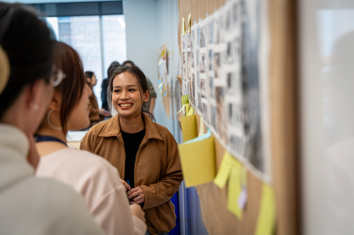 A student smiles at a group of her peers while standing by a board with photos and yellow sticky notes on it. 