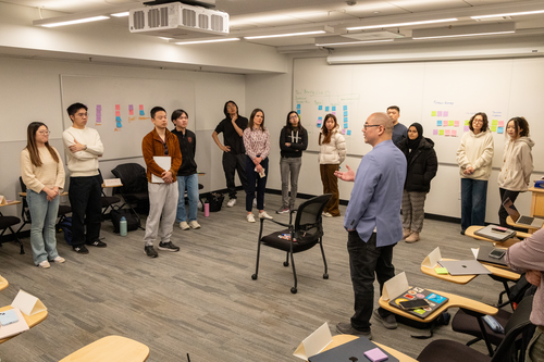 A classroom of students stands in a horseshoe around the classroom with Professor Isotani in front. An empty chair is in the middle of the room, and the white boards on the two walls are full of multi-colored post-it notes. 