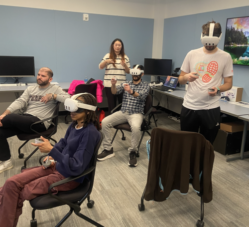 Three people wear VR headsets on their heads and aim controllers. Another person in a Penn sweatshirt is seated with them, and a teacher stands, not wearing a headset, giving direction. 