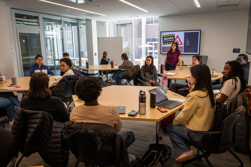 Ameena stands at the front of a classroom in front of a screen with a slide on it about checking in on their reports in progress. Students sit clustered around four large tables engaged in discussion. 