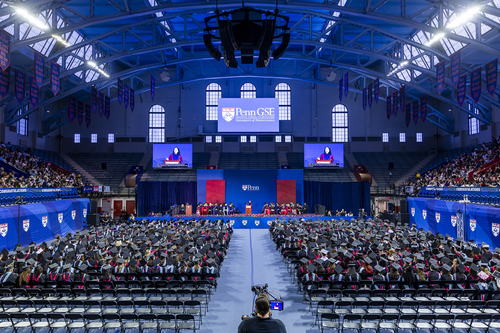 Wide interior view of a large commencement ceremony in an indoor arena, with rows of graduates seated facing a stage, large video screens, and a backdrop displaying ‘Penn GSE.’