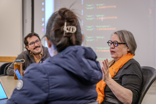 Yasmin Kafai (right) gestures while instructing students around a seminar table in front of a screen displaying computer code.