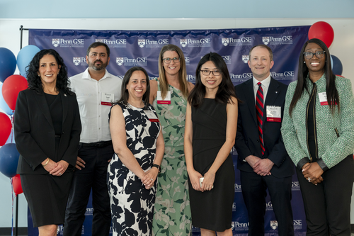Dean Strunk and six of this year's Education Alumni Award winners pose in front of a Penn GSE background and blue and red balloons. 