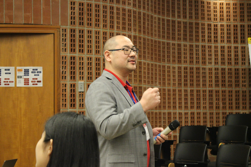 Seiji Isotani stands in the audience engaging with someone offscreen and holding a microphone