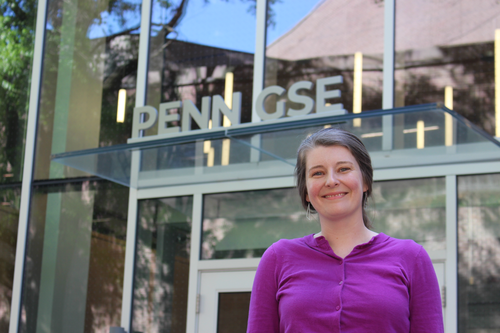 Amy wears a purple shirt and smiles in front of the glass front of the Penn GSE building. The Penn GSE sign is visible behind her. 
