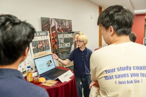 A person gestures to a laptop with their venture on it. A sign in front reads Lingriser. A person looking on, whose back is to the camera, is wearing a shirt advertising Ask Sia. 