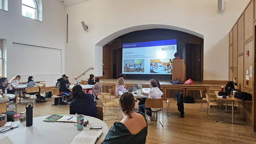 A teacher presents onstage with a screen that reads "Summer Institute: Seting the Context" to a room of teachers seated at round tables in a sunny room. 