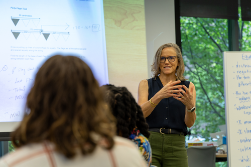 Caroline Ebby gestures with her hands in front of a screen showing math equations. The back of teachers heads, sitting in her class, are visible. 