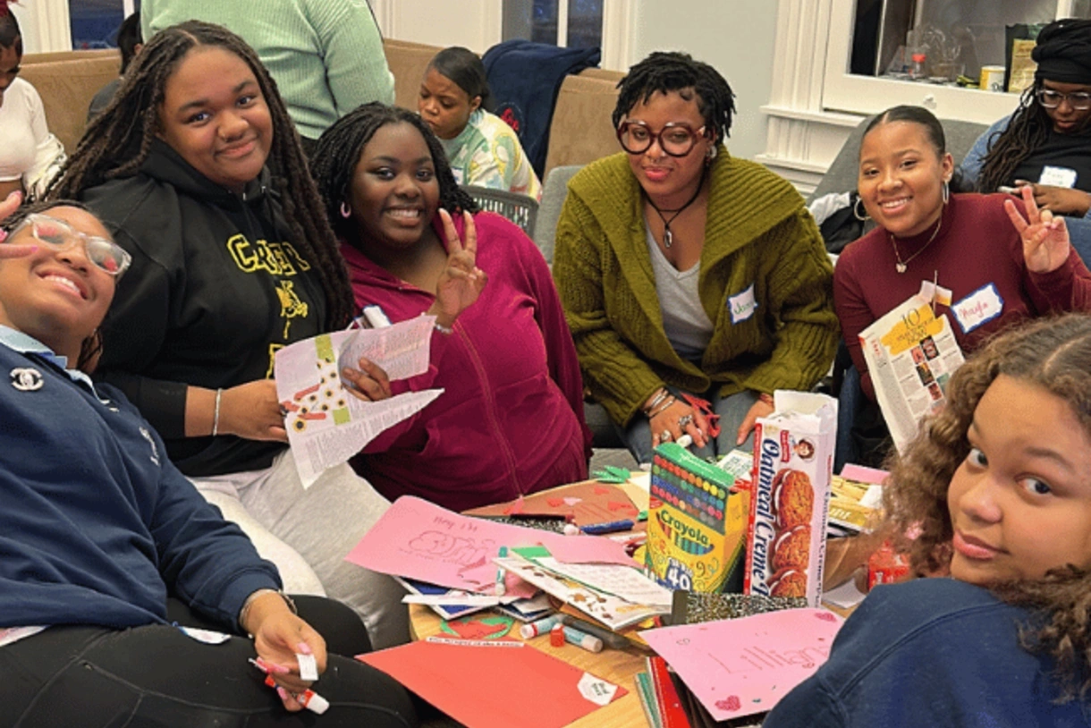 Image of local students–primarily 6 black girls sitting around a table with art supplies.