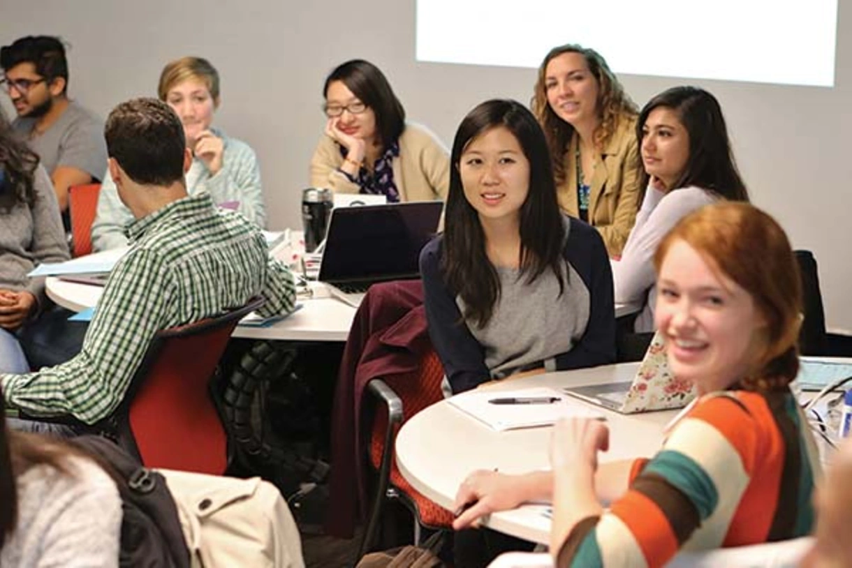 Graduate students sitting at round tables, interacting with each other or looking ahead, with smiles on their faces.