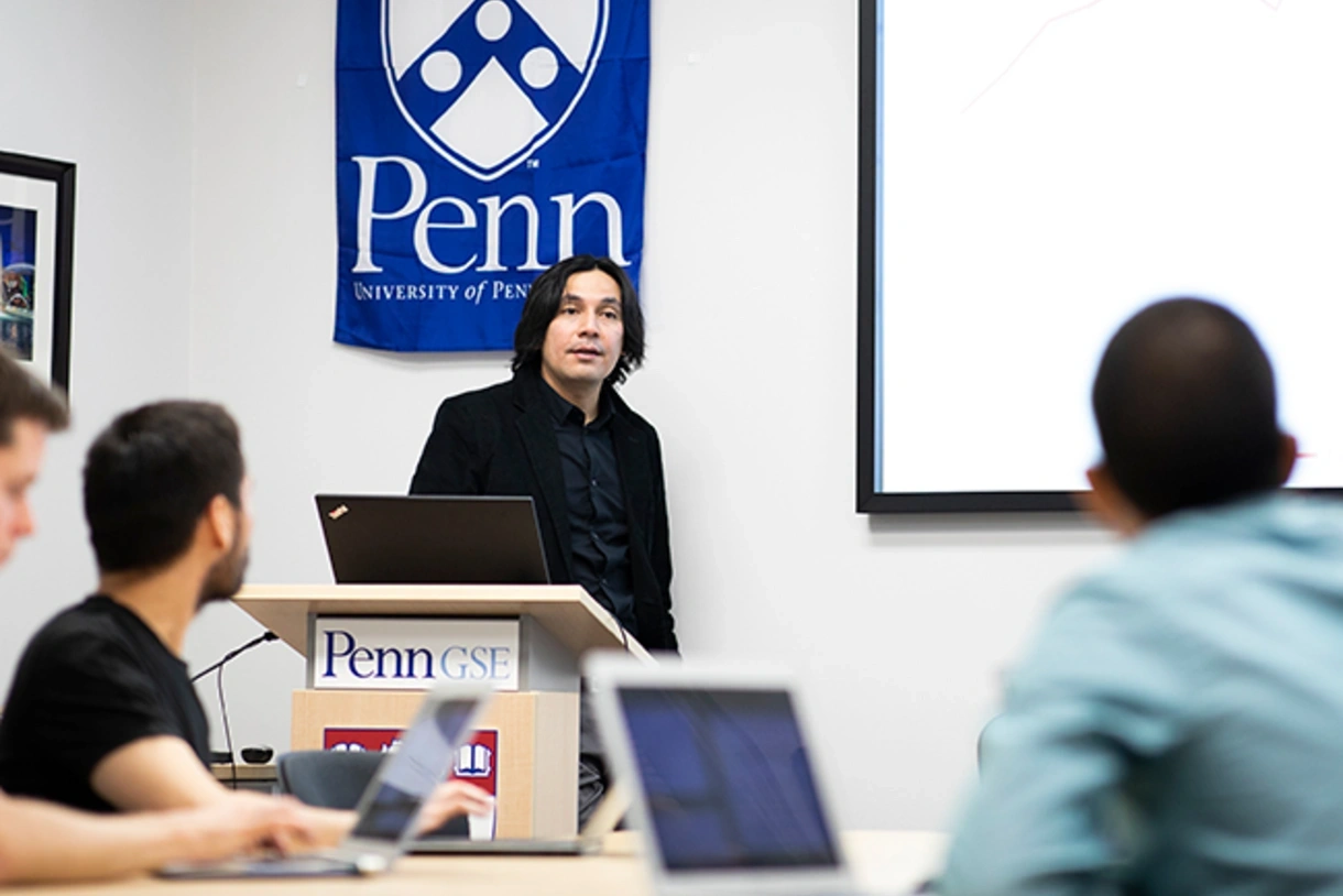 A professor wearing a suit speaks to students in a classroom. A blue Penn banner is behind him, and a Penn GSE logo is on the podium where he stands.