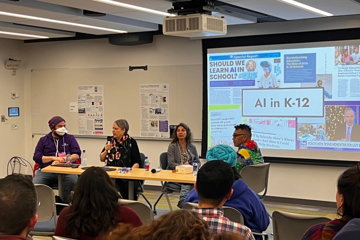 Four people sit in a row at a long table in front of an audience with a slide that says “AI in K–12” on a screen behind them.