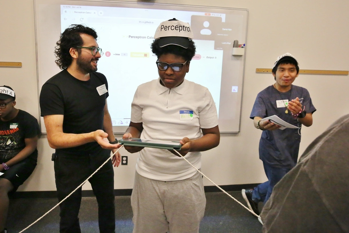 Luis running a workshop with students, helping a student wearing a hat that reads "Perceptron" and holding an iPad and strings that are connected to several of his classmates. 