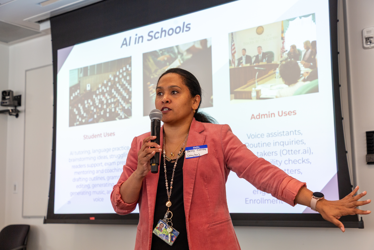 Betty Chandy holds a microphone, presenting in front of a screen that reads "AI in Schools"