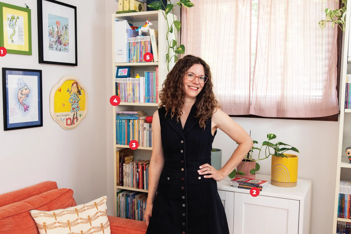 Katie Carella wears glasses and smiles with her hands on her hips inside her colorful office that is full of books
