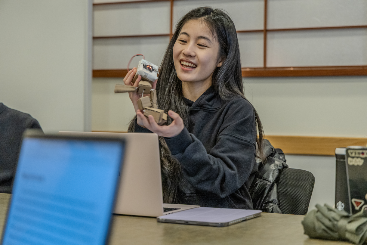 A student seated at a seminar table holds up a small handmade robot