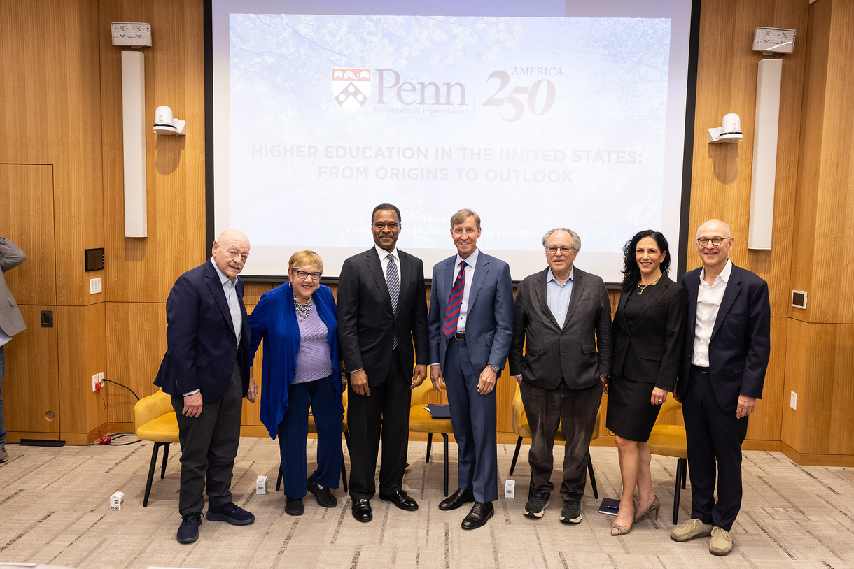 Seven university leaders dressed in suits stand in a line smiling in front of a screen that reads Penn | America 250