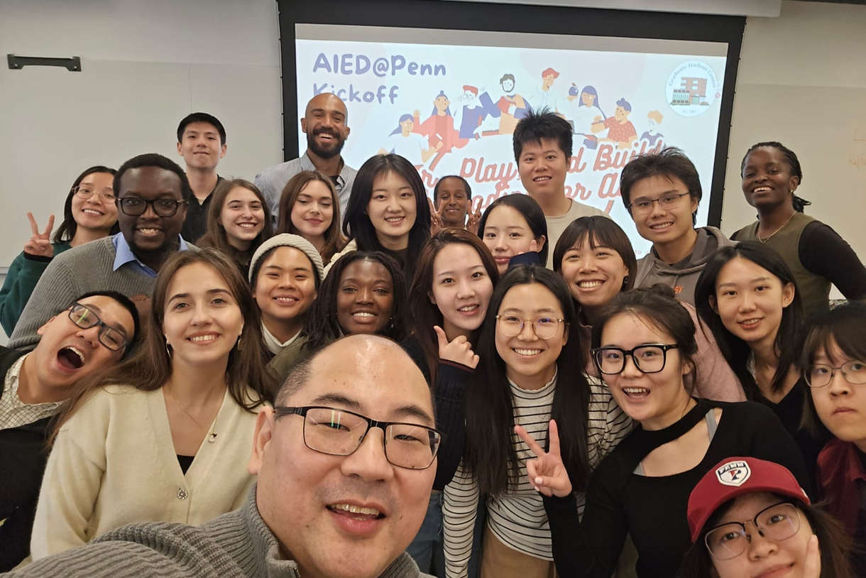 A large group of students take a selfie with Seiji Isotani at the front and a screen with AIED@Penn visible on it