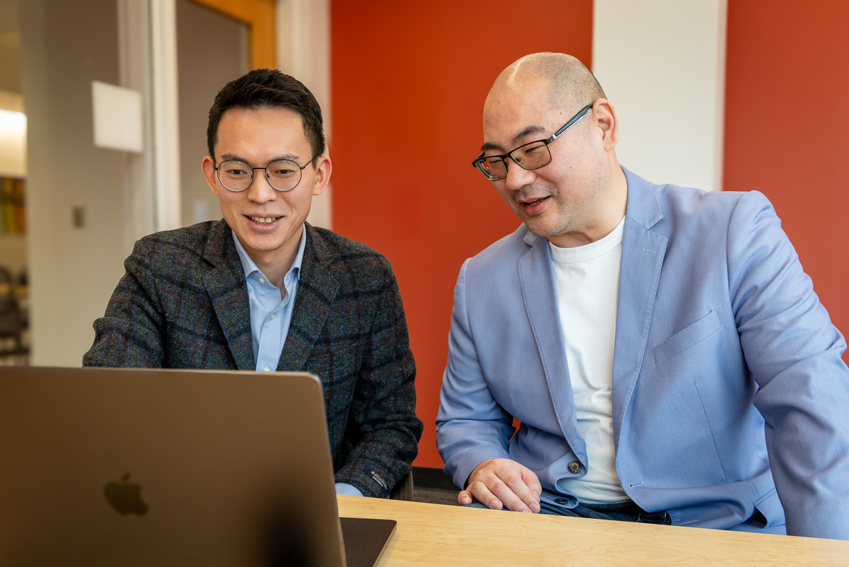 Bodong Chen, left, and Seiji Isotani sit at a table side-by-side in sport coats reading from the same laptop.