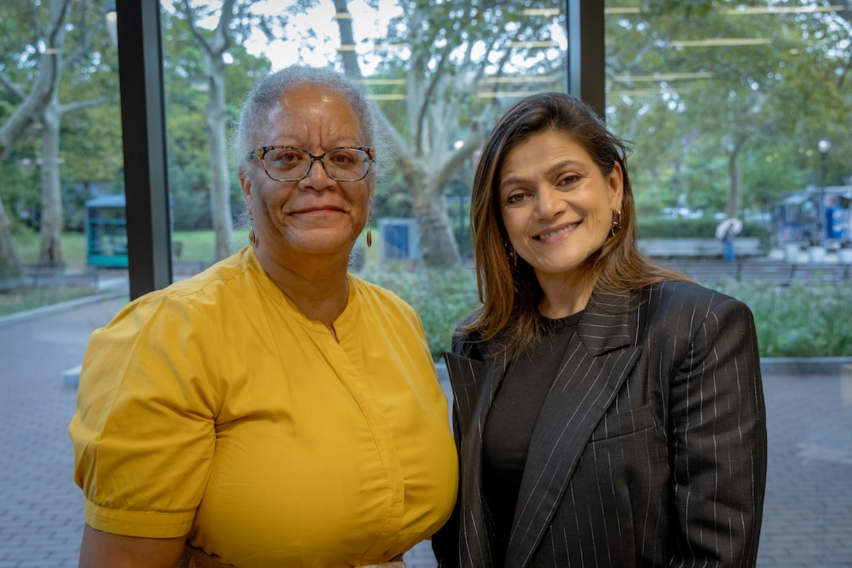 Marsha and Neerja stand smiling in front of a wall of windows
