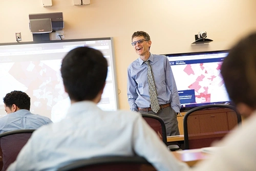A professor wearing a blue shirt and patterned tie, smiles and addresses students in a classroom. Behind him are screens containing digital media.