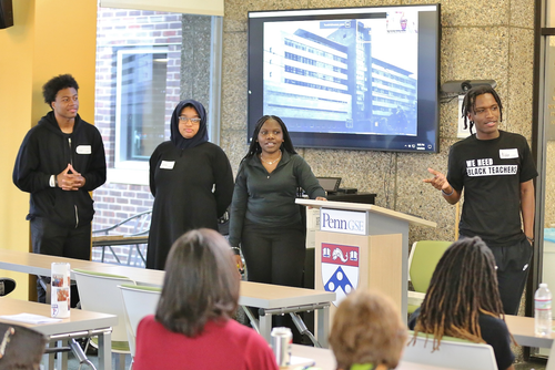 Four students, including one wearing a t-shirt that says "We need Black teachers," present at a podium with a Penn GSE logo and crest on it in front of a room of people seated at tables. 