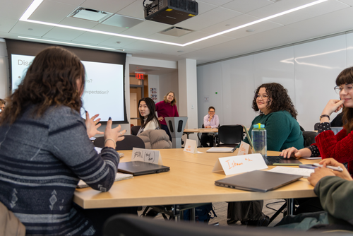 Viewed from the back of the classroom, we see students speaking and gesturing with their hands and between them the two professors are visible at the front of the room. 