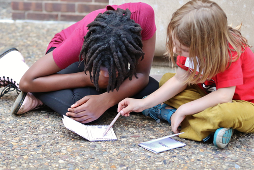 Two kids huddled over a notebook writing outdoors.