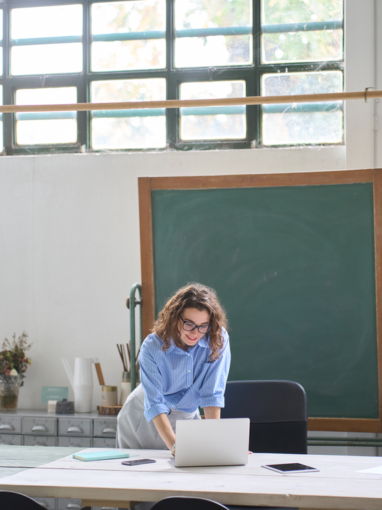 A female teacher in a classroom hovering over a desk looking at a laptop.