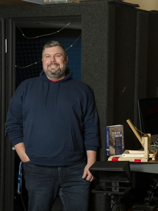 Trey stands in front of a recording booth and next to a 3D printer and a table full of books, awards, and items made in the makerspace.