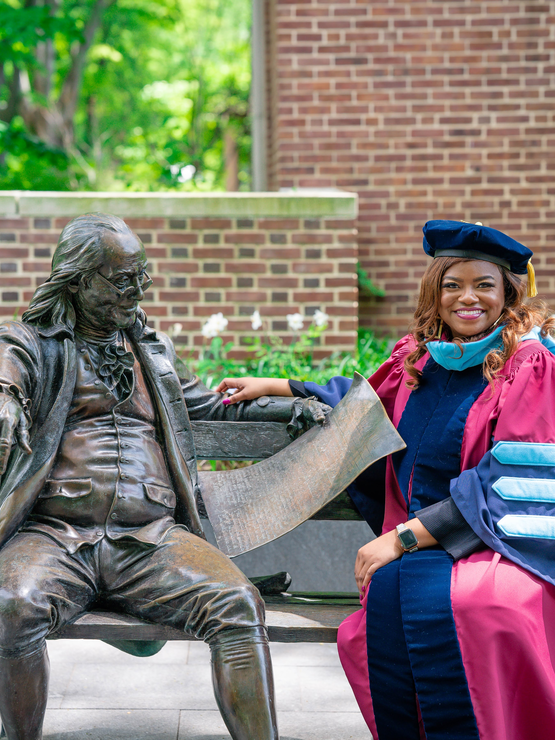 Kimberlin wears her red-and-blue doctoral robe and cap, sitting on a bench with the Ben Franklin statue near Locust Walk. 
