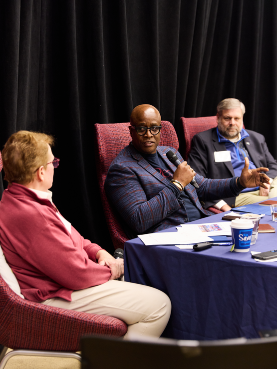 Roger Ward, seated in between Karen Weaver and Jeff Doggett, holds a microphone and talks